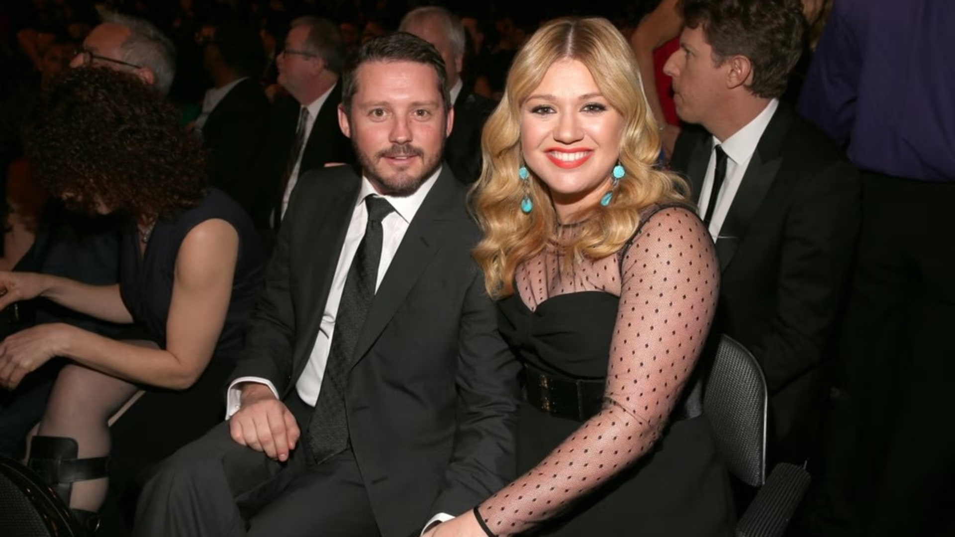 Brandon Blackstock and Kelly Clarkson are seated together at an awards event, smiling toward the camera among guests in a dim venue