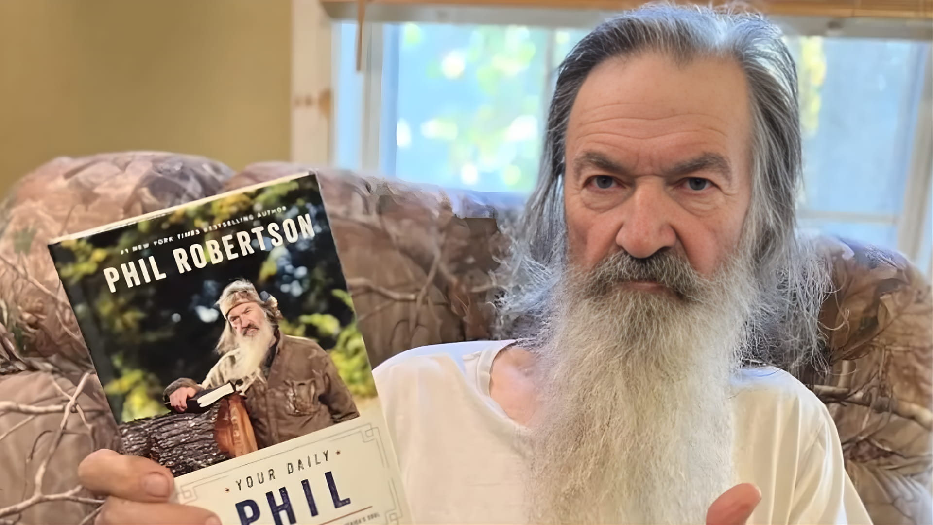 Phil Robertson holding his book while sitting indoors, with a long beard and serious expression, reflecting his public image