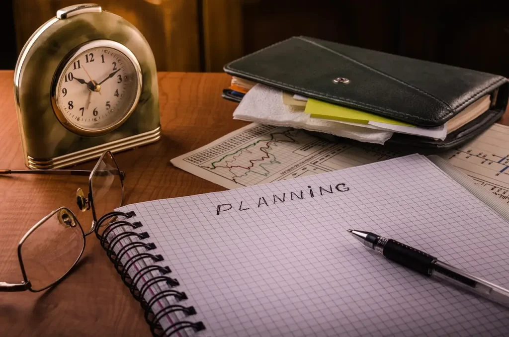 Notebook with "Planning" on wooden desk alongside clock, glasses, graphs, and planner