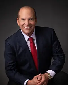Man in dark suit with red tie against gray background, smiling confidently in portrait setting