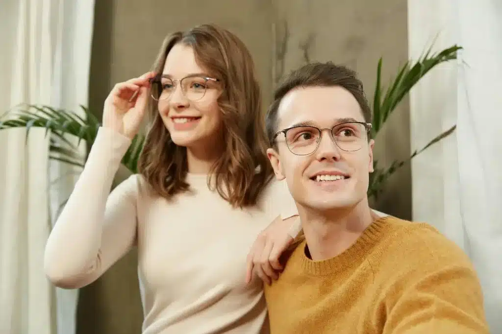 Two people smiling and wearing glasses in a cozy living room setting with plants