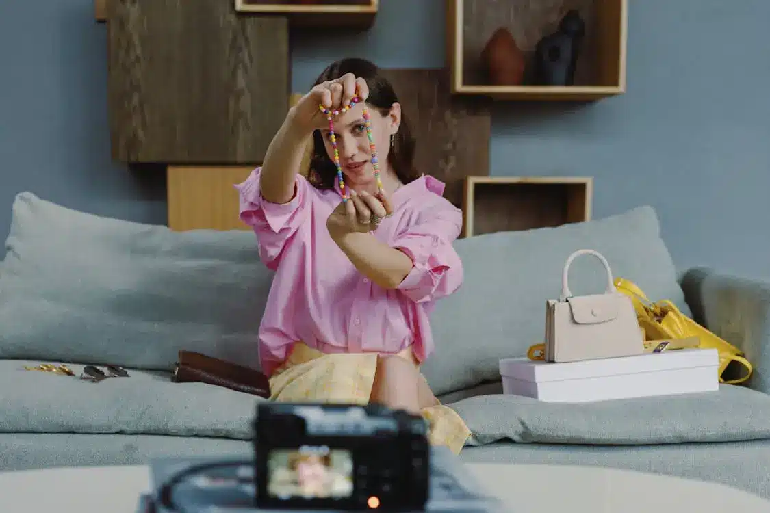 Person in pink shirt showcasing necklace on couch with camera and handbags nearby