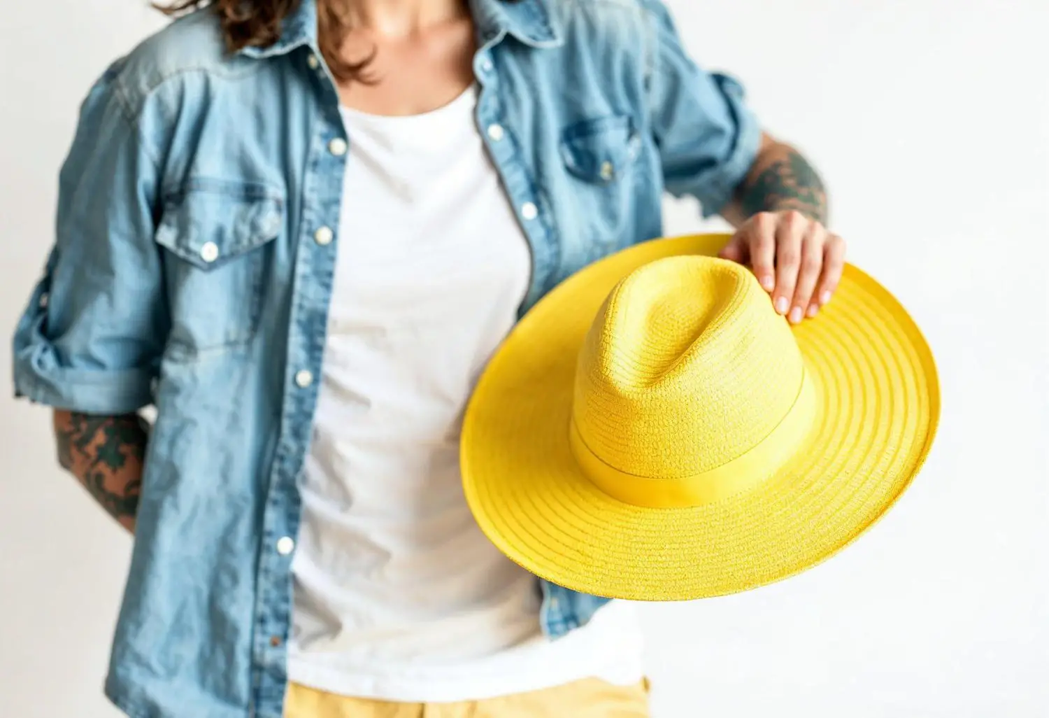 Person in denim shirt holding yellow straw hat against white background