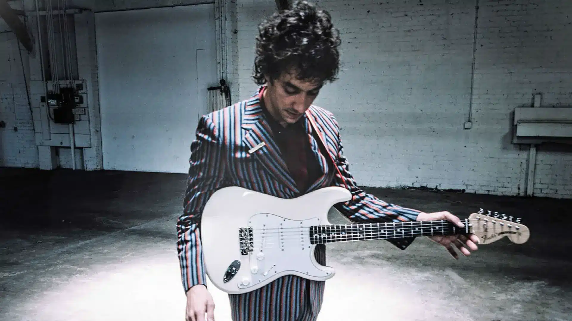 Albert Hammond Jr., guitarist of The Strokes, holding a white Fender Stratocaster in an empty industrial space