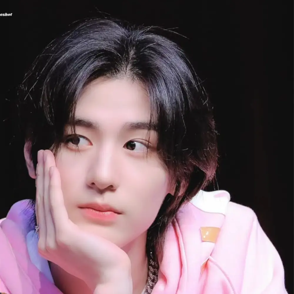 BOYNEXTDOOR member resting their face on hand, wearing a pink hoodie and silver bracelet, looking thoughtfully to the side.