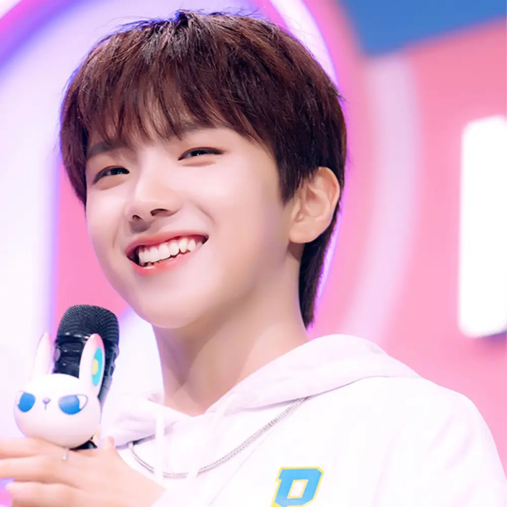 BOYNEXTDOOR member smiling while holding a microphone with a small white toy, wearing a white hoodie on a colorful stage.