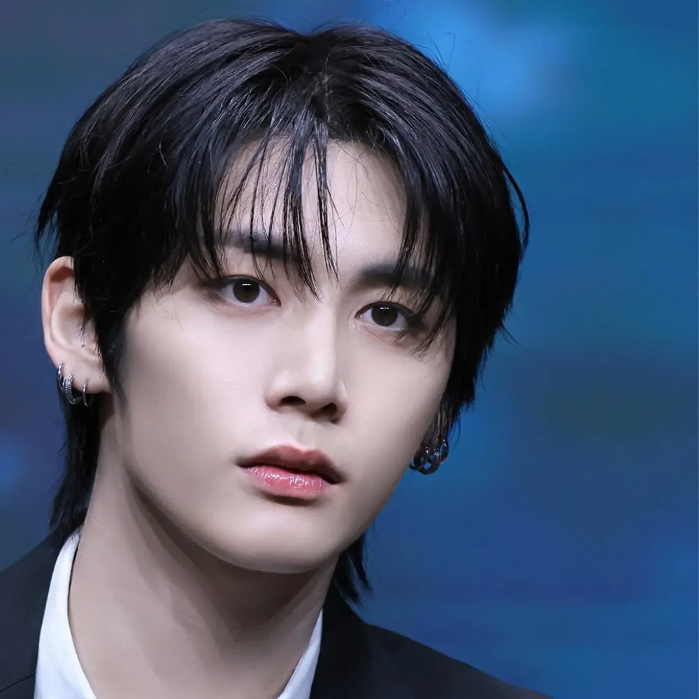 BOYNEXTDOOR member wearing a black suit, white shirt, and black tie, looking forward with dark hair and earrings.