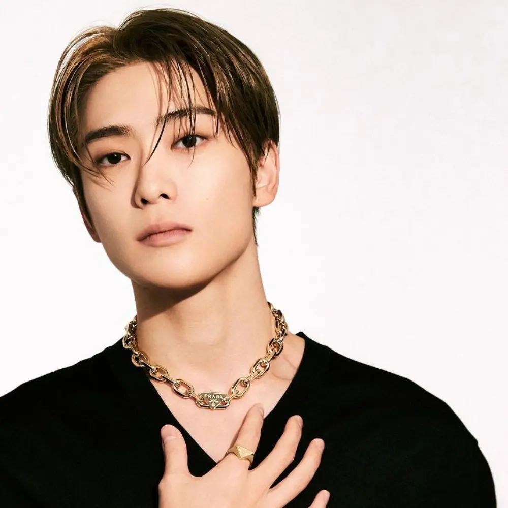 BOYNEXTDOOR member wearing black Prada top and gold chain necklace, posing with hand on chest against a white background