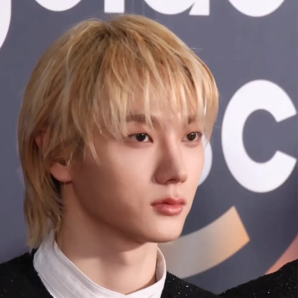 BOYNEXTDOOR member with blonde hair wearing a black glitter jacket and white shirt, looking to the side against a blurred backdrop.