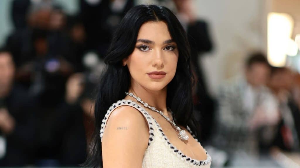 Dua Lipa posing at a red carpet event wearing a white dress and diamond necklace