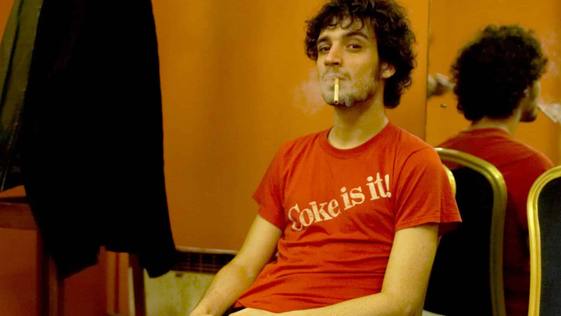 Fabrizio Moretti, drummer of The Strokes, sitting in a room with an orange wall, wearing a red Coke is it! t-shirt and smoking a cigarette.