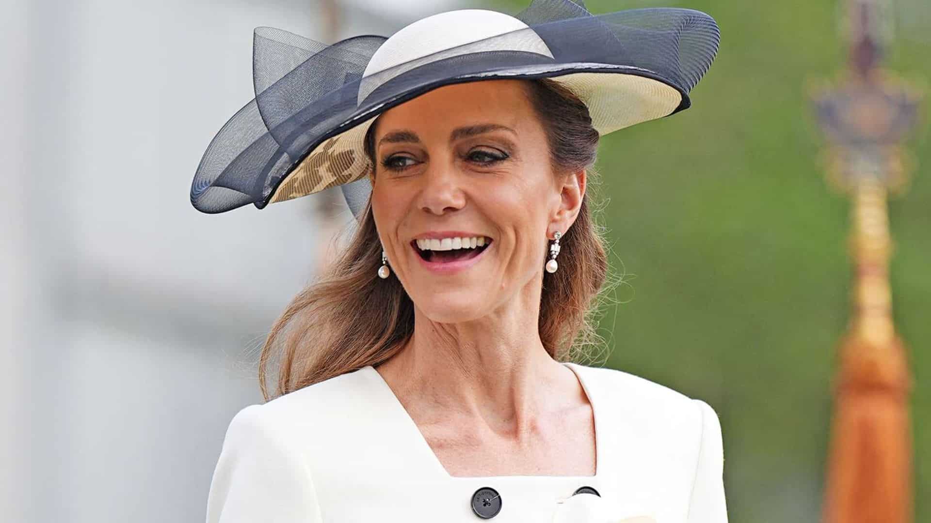 Kate Middleton smiling, wearing a white outfit with a black and white hat