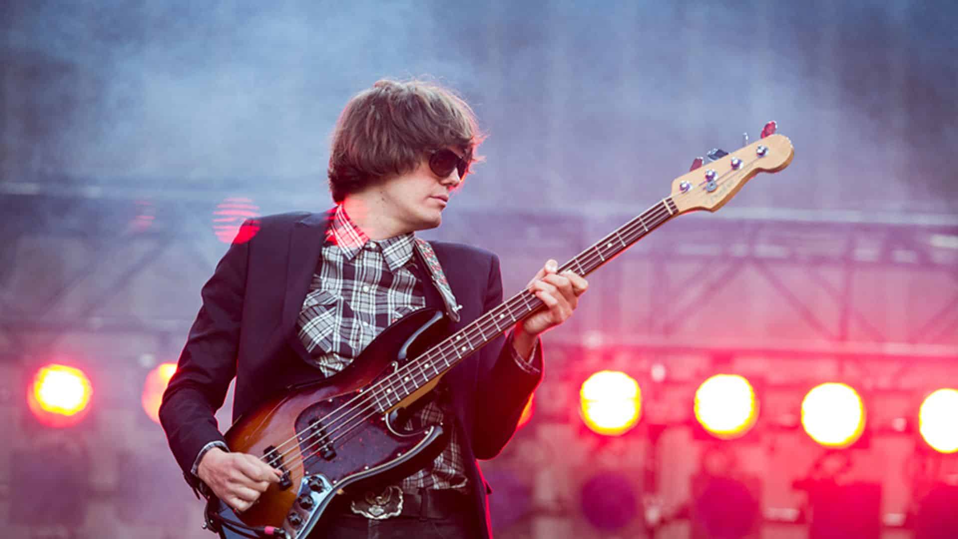 Nikolai Fraiture, bassist of The Strokes, playing a sunburst bass guitar on stage with bright red stage lights behind him