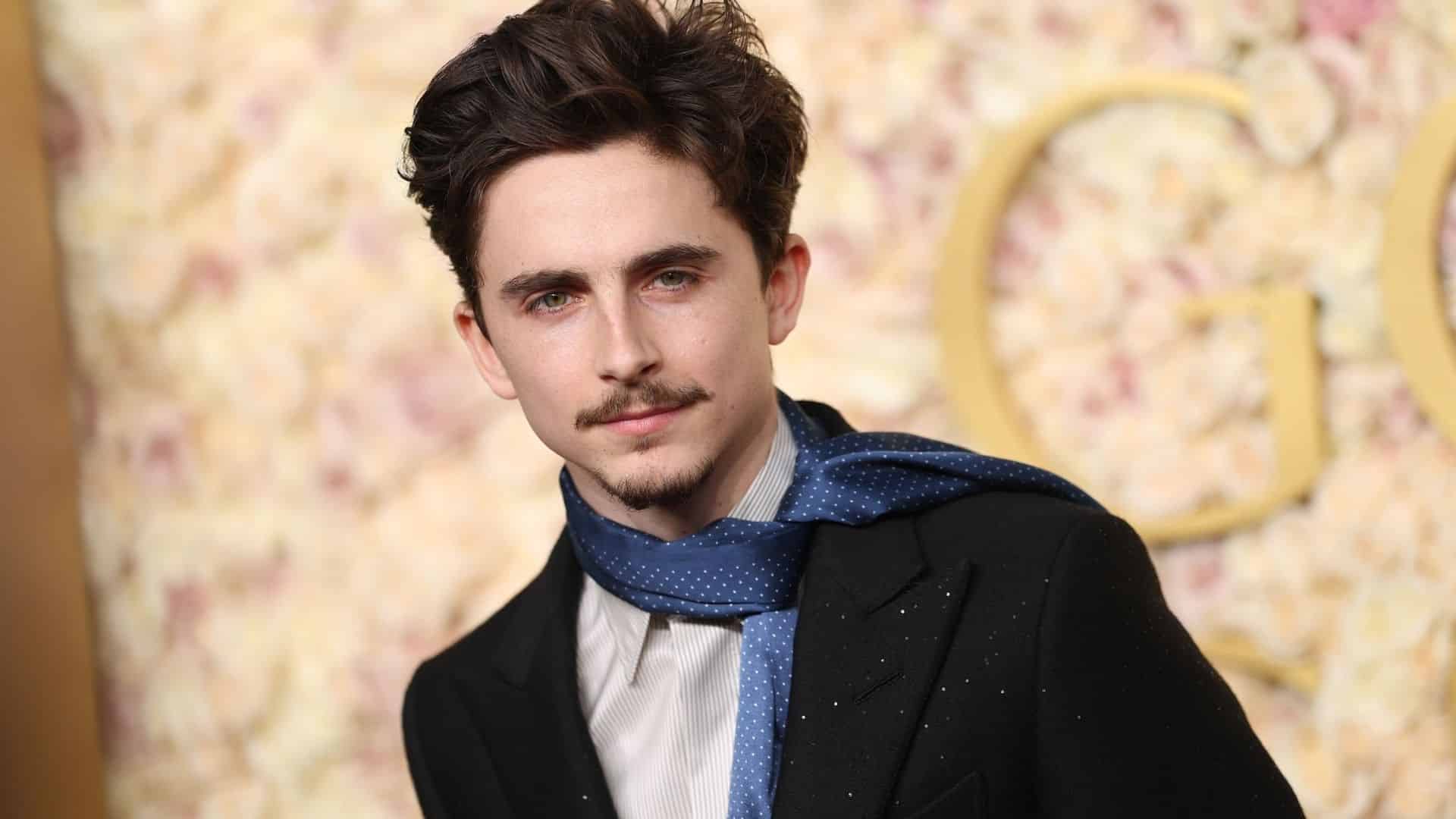 Timothée Chalamet posing in a black suit and blue scarf at a formal celebrity event.