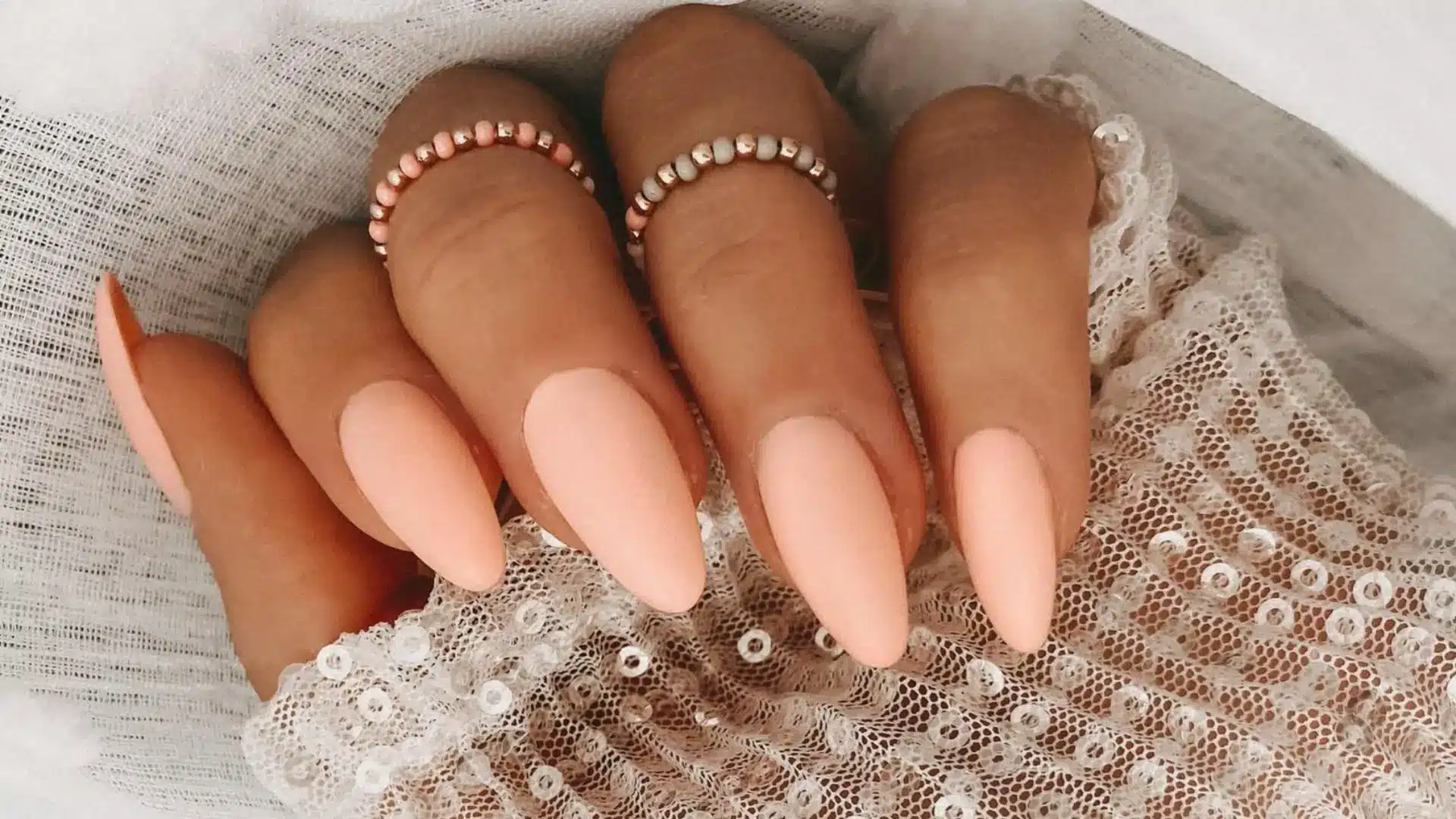 A hand with Almond-Shaped Peach Nails, paired with delicate rings, set against a soft, textured background