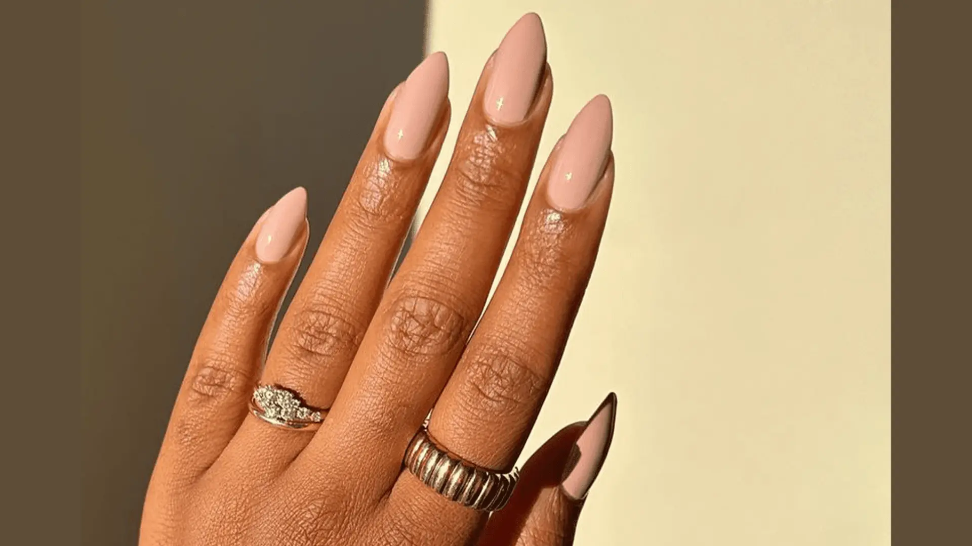 A hand with natural beige nails, adorned with delicate rings, against a soft background