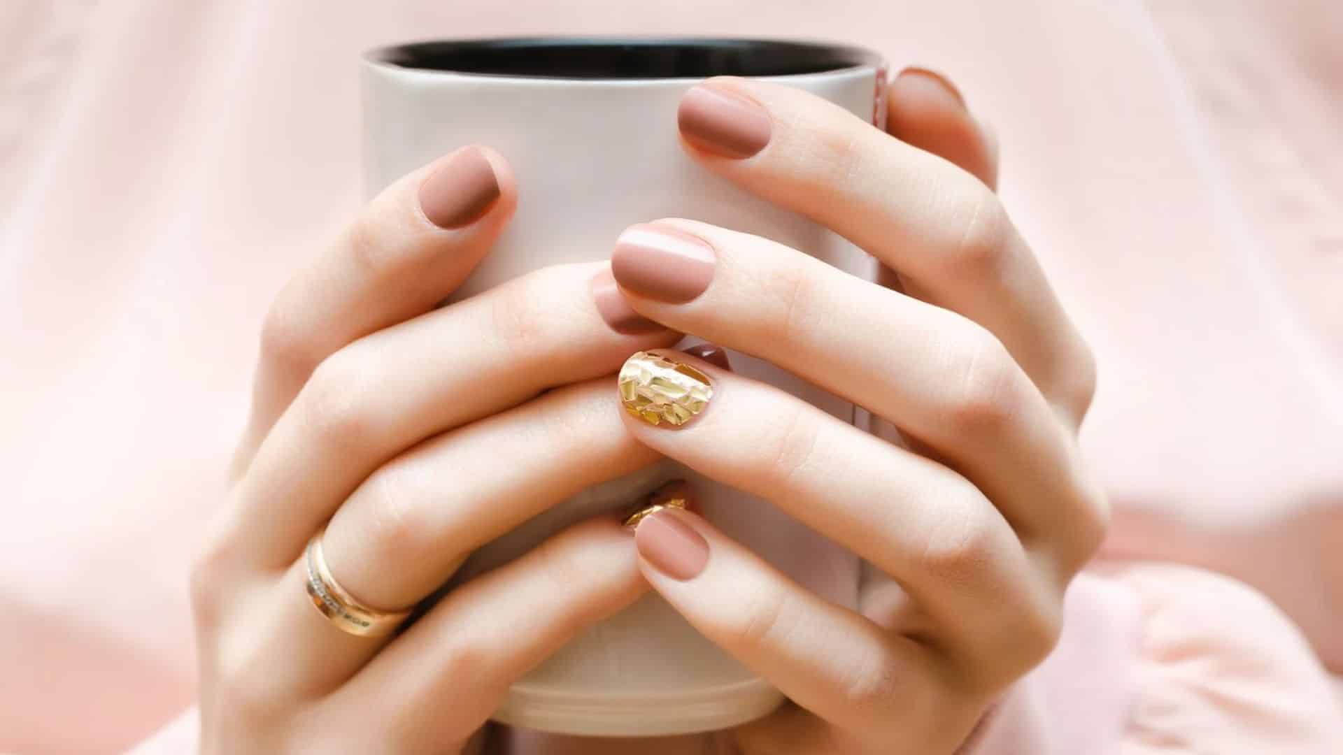 A pair of hands with Nude Matte Nails, featuring a unique gold-accented nail, holding a cup