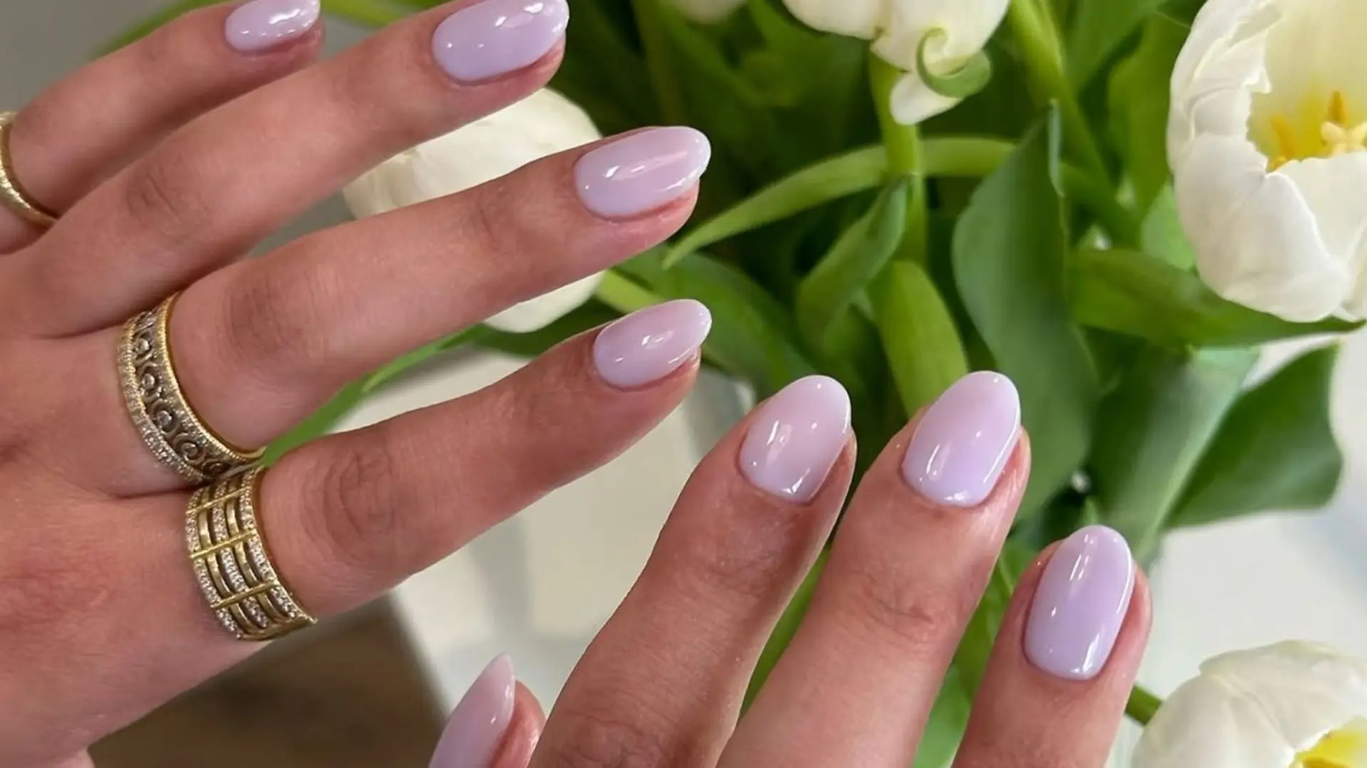 A pair of hands with soft pastel nails, accessorized with gold rings, in front of a bouquet