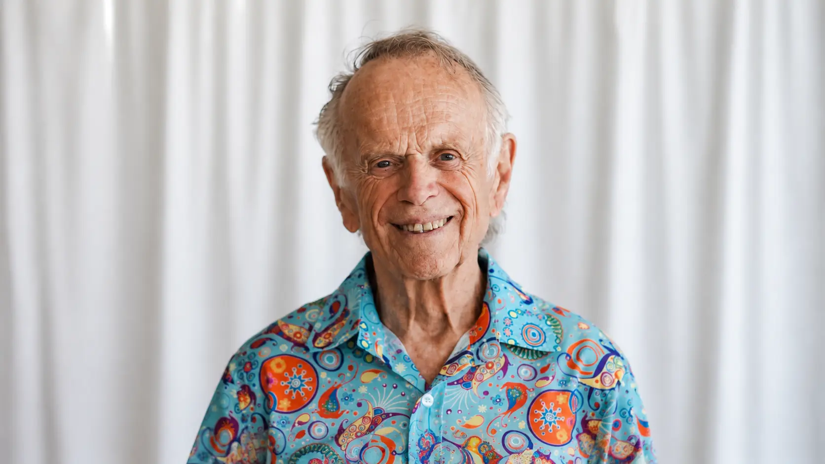 Elderly man smiling warmly at the camera, wearing a colorful patterned shirt against a simple light studio background.