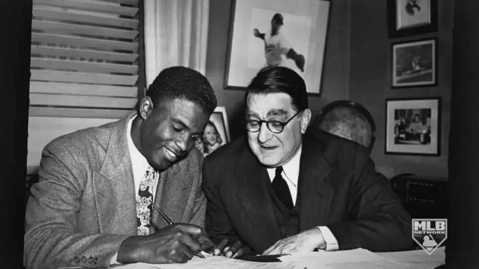 Black-and-white photo of two men in suits signing baseball-related documents at a desk, with framed sports photos in the background