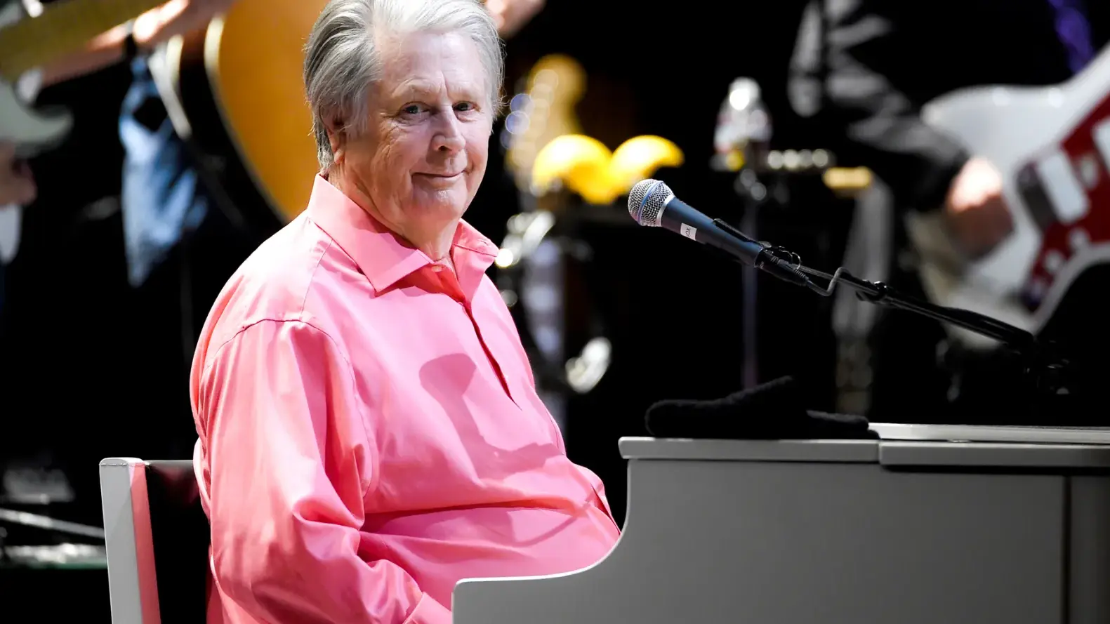 Brian Wilson of the Beach Boys playing piano on stage, wearing a pink shirt with band members and instruments in the background.