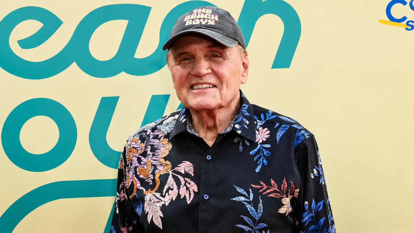 Older man in floral shirt and Beach Boys cap smiles at event with teal and yellow backdrop.