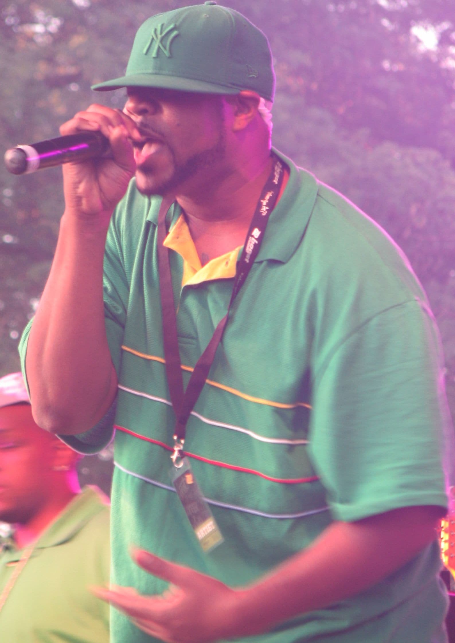 Capadonna from Wu-Tang Clan performing on stage, wearing a green cap and striped green polo shirt, holding a microphone