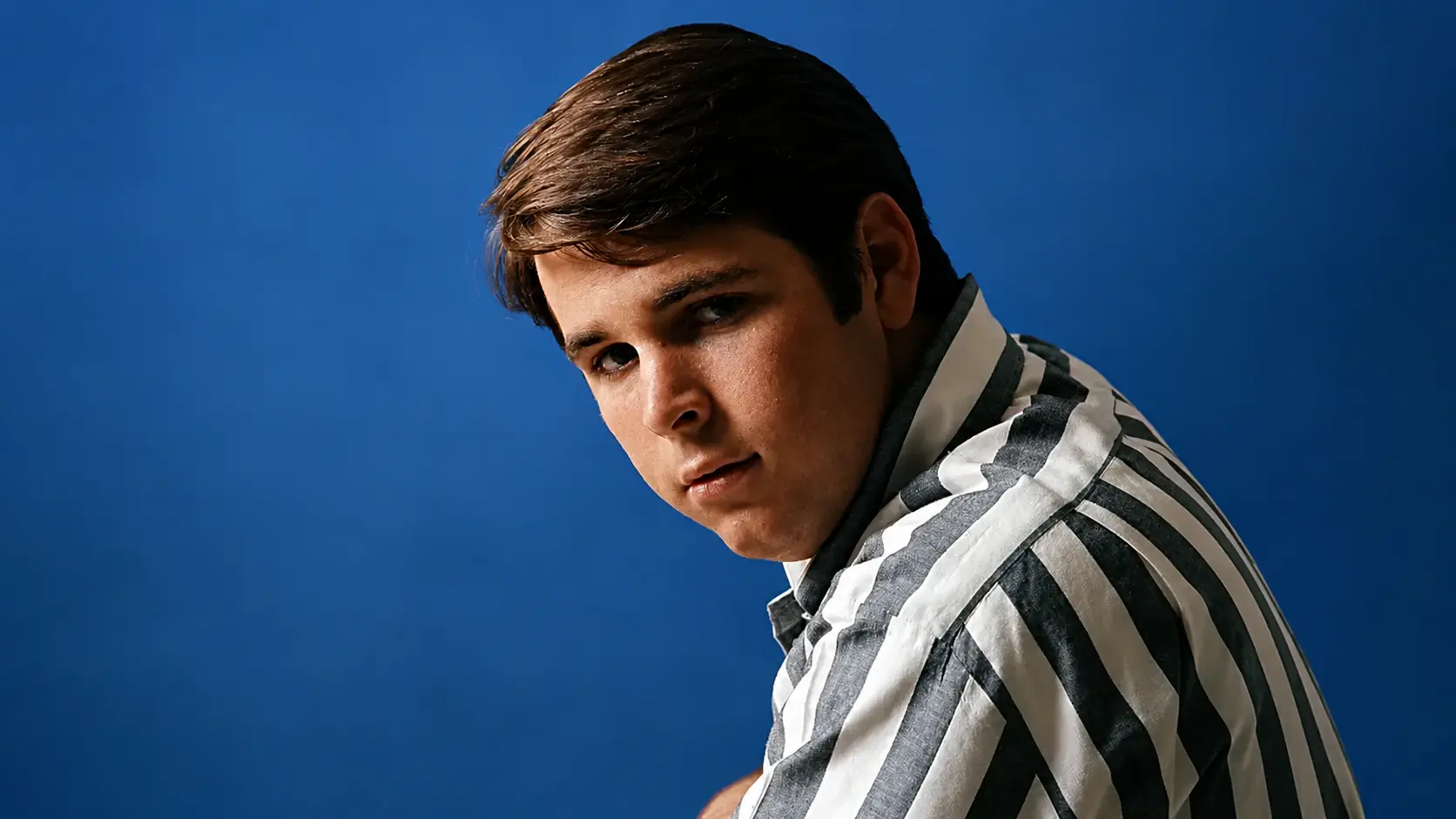 Young man in a striped shirt looks sideways at the camera against a solid blue background, vintage portrait style.