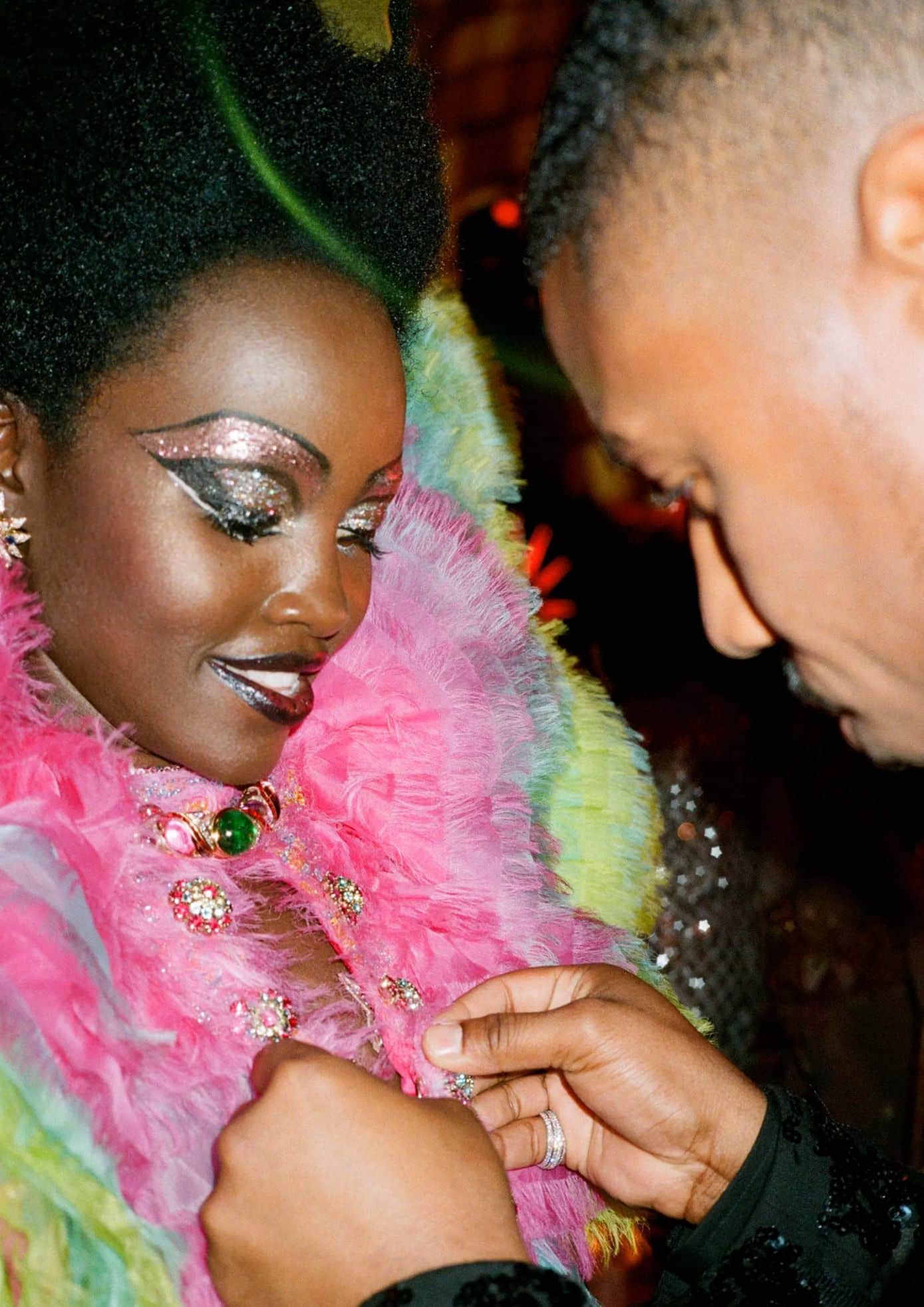 Close-up of a woman in pink feathered outfit with sparkling makeup, as a man adjusts her jewelry at the 2019 Met Gala. (1)