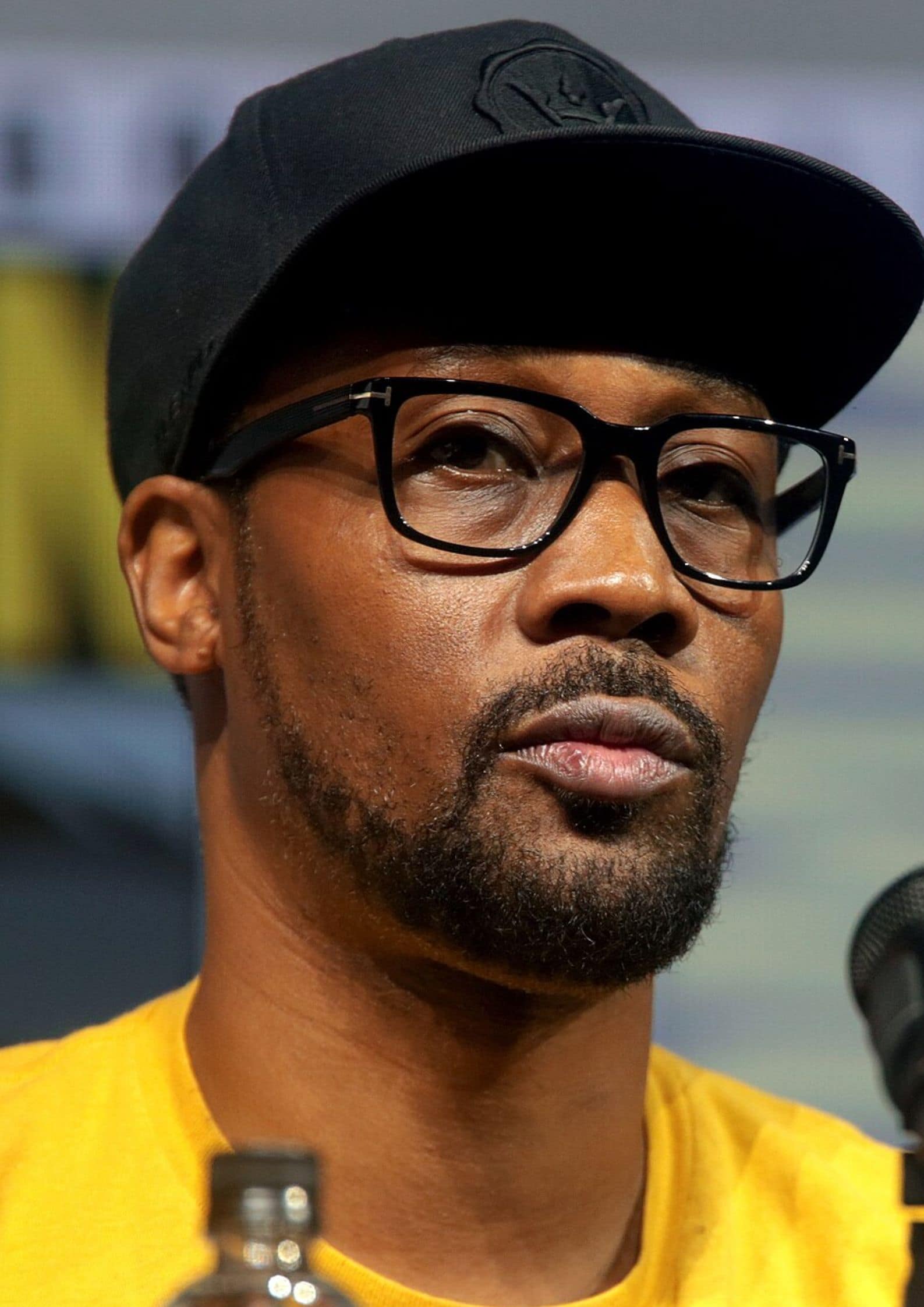 Close-up of RZA from Wu-Tang Clan wearing a black cap, glasses, and yellow shirt speaking at a microphone.