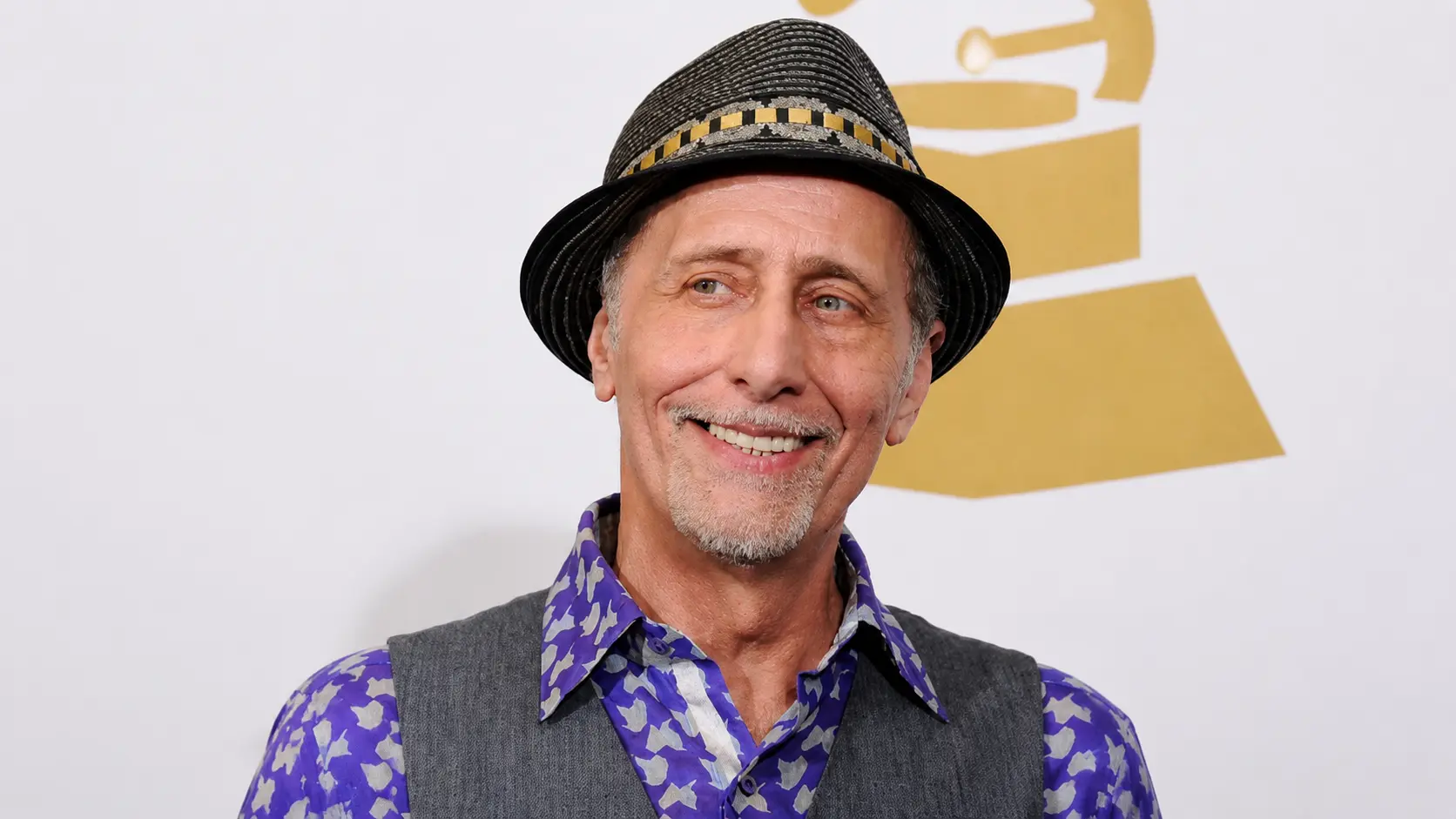 Older man smiling on a red carpet, wearing a patterned purple shirt, gray vest, and black checkered fedora.