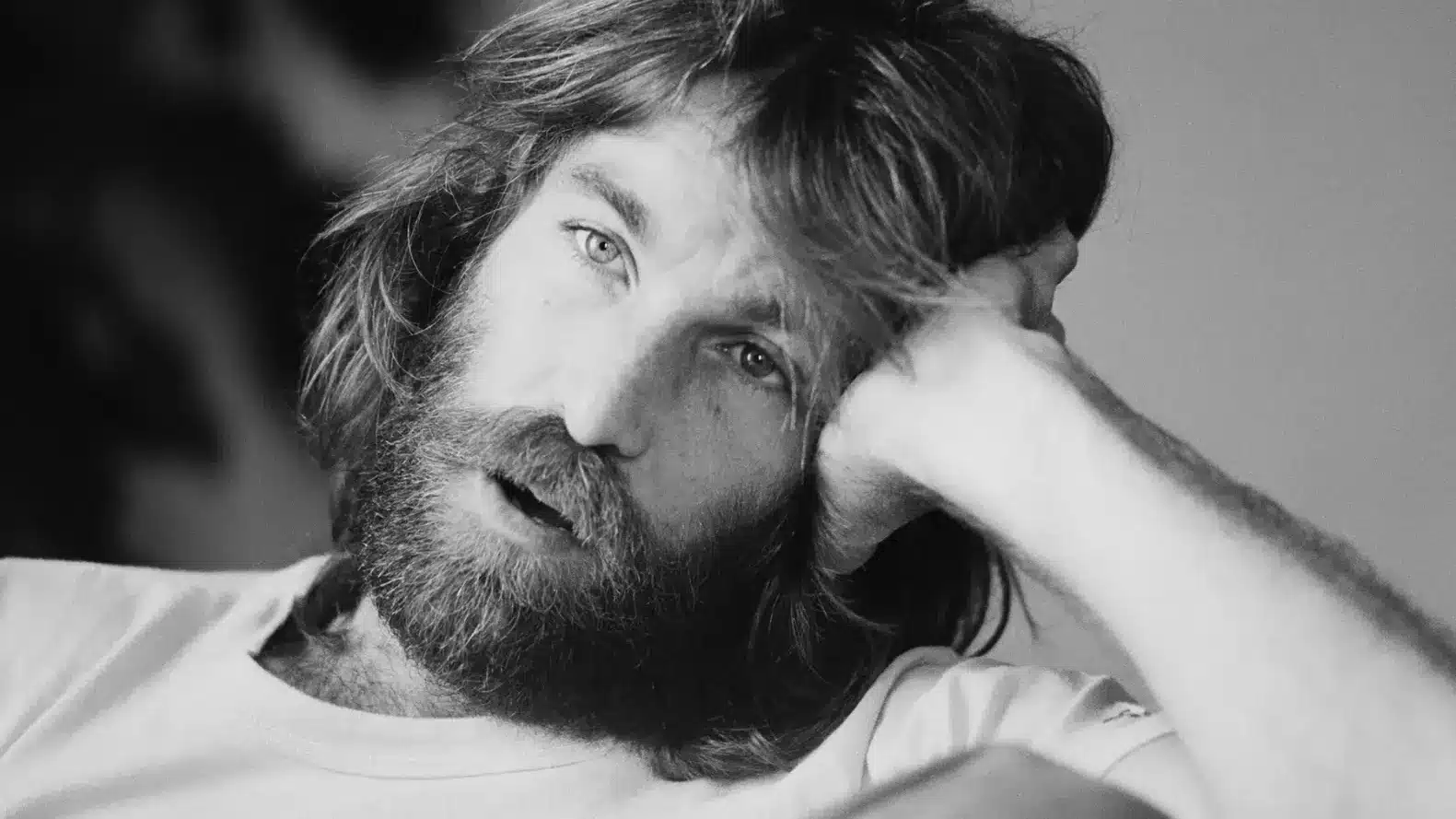 Close-up black-and-white photo of Dennis Wilson of the Beach Boys with long hair and beard, resting his head on his hand.