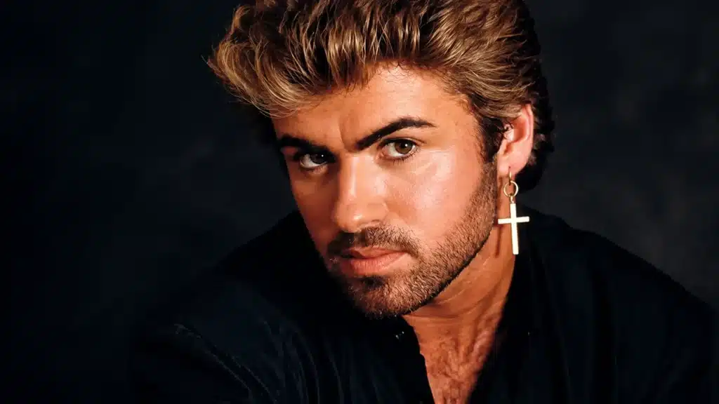 George Michael close-up portrait wearing a cross earring with styled hair and a dark background.