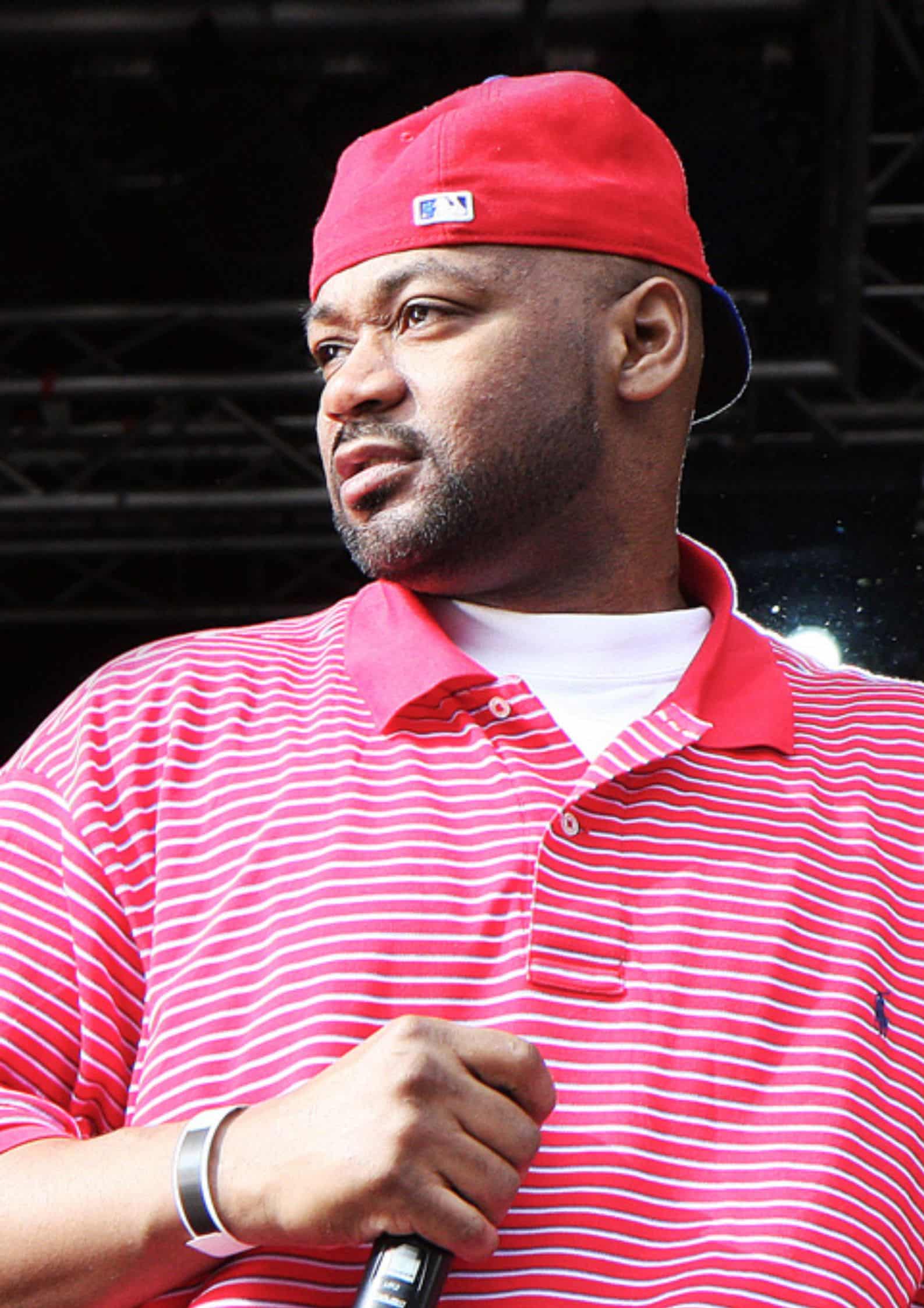 Ghostface Killah (Dennis Coles) from Wu-Tang Clan wearing a red cap and red striped polo shirt, holding a microphone on stage.
