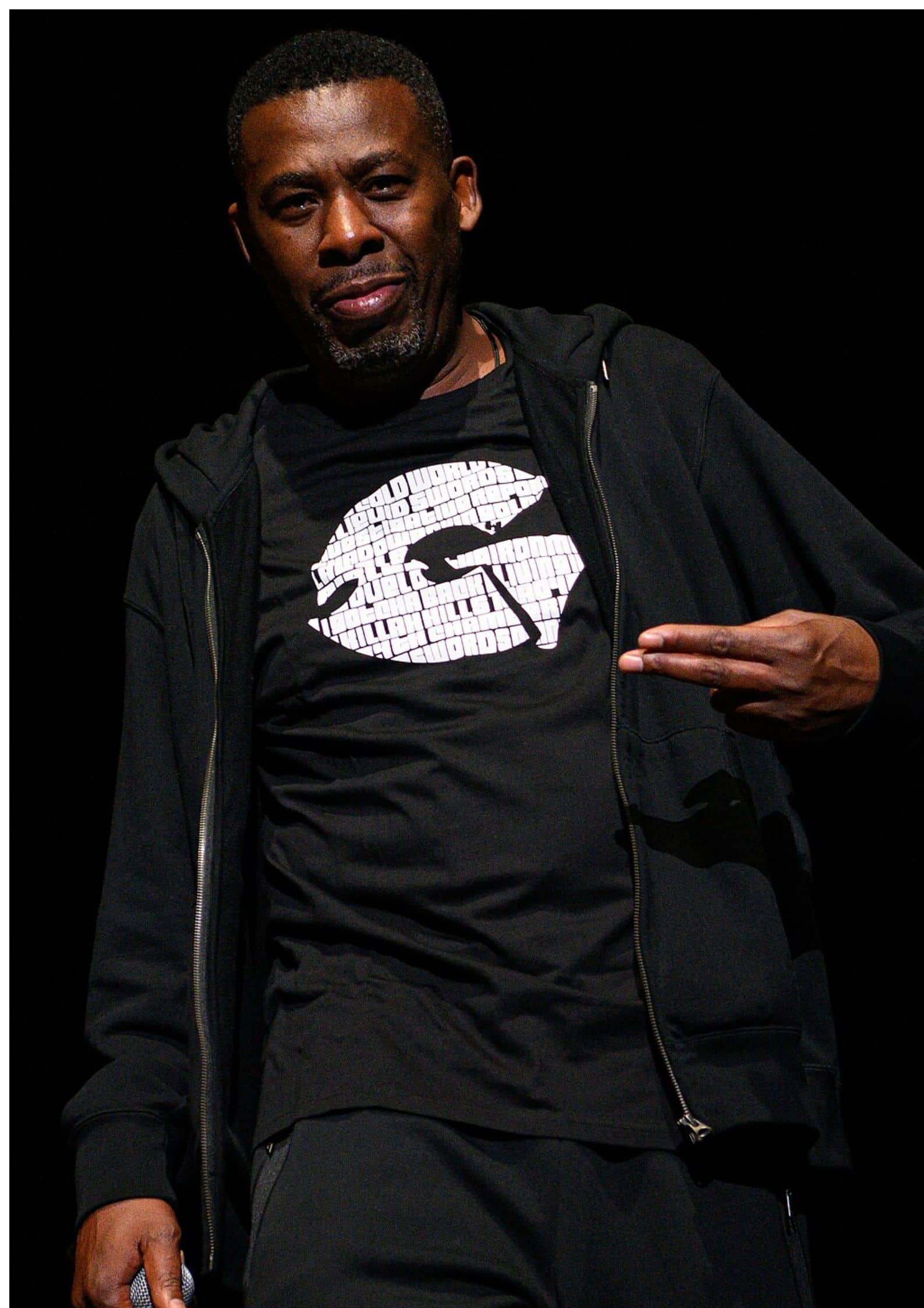 GZA from Wu-Tang Clan wearing a black hoodie and t-shirt, posing with hand gesture against a dark background.