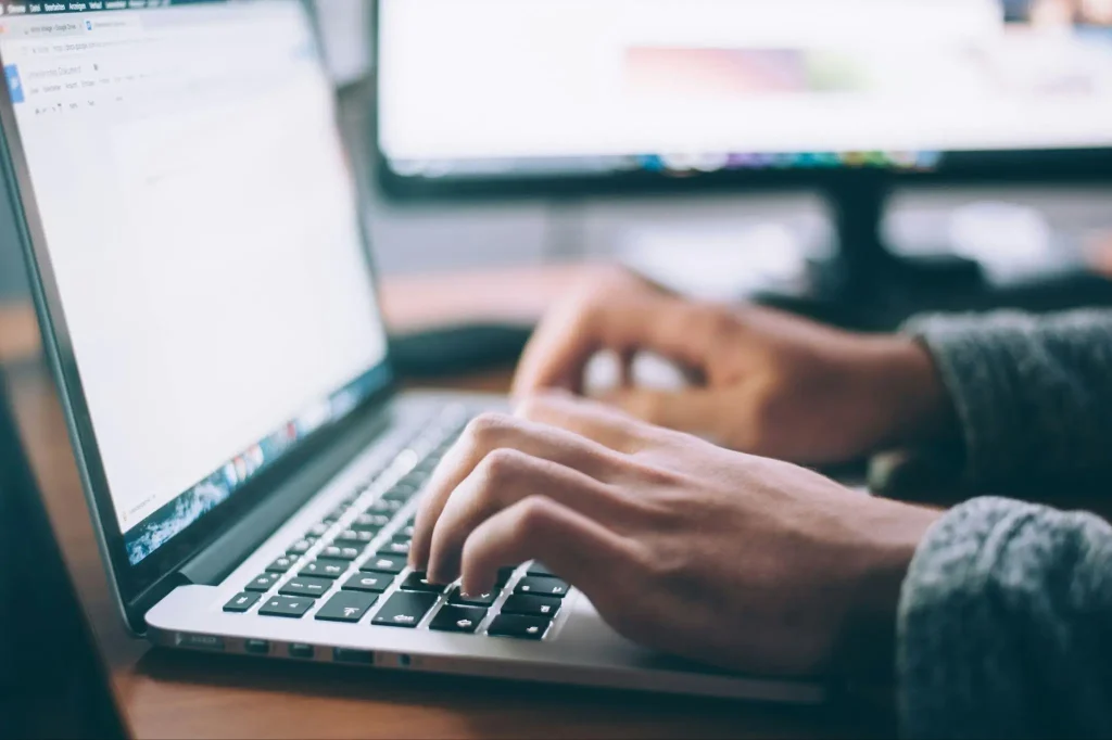 Hands typing on a laptop keyboard with a blurred monitor in the background