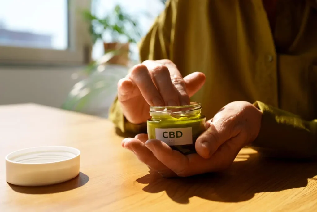 Hands holding CBD oil jar on wooden table in sunlit indoor space