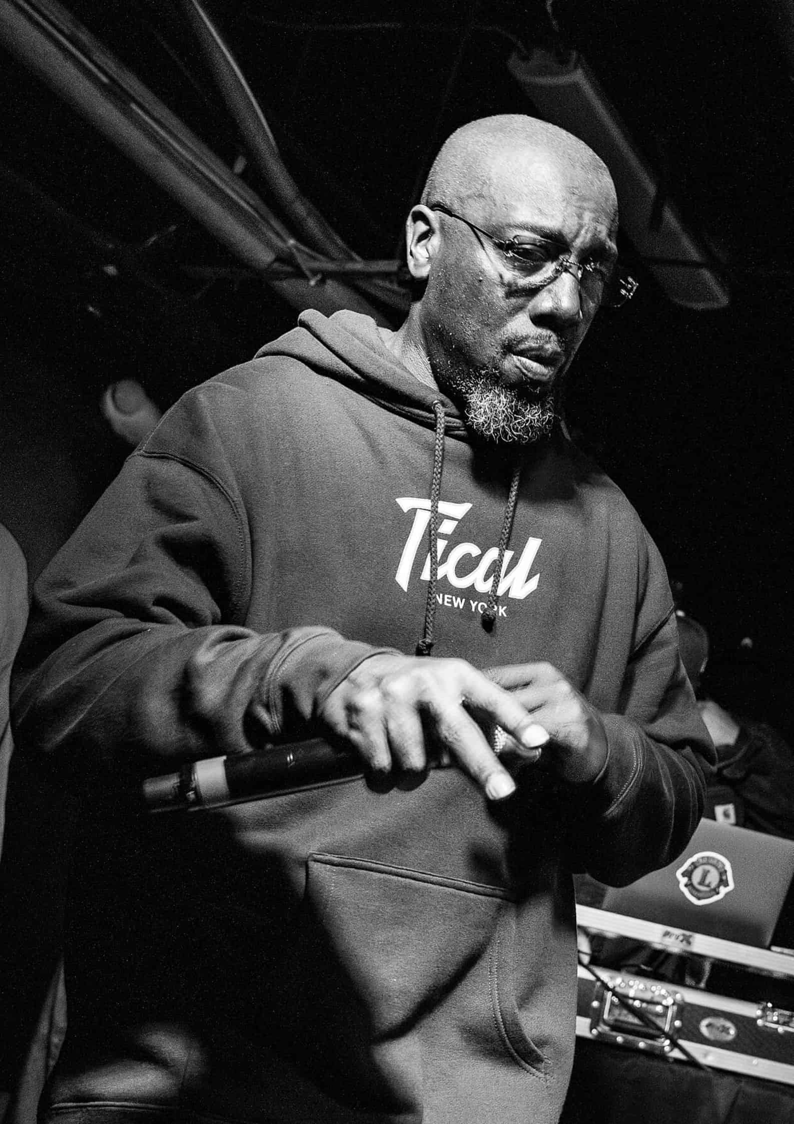 Inspectah Deck (Jason Hunter) from Wu-Tang Clan wearing glasses and a hoodie, performing on stage in black-and-white photo.