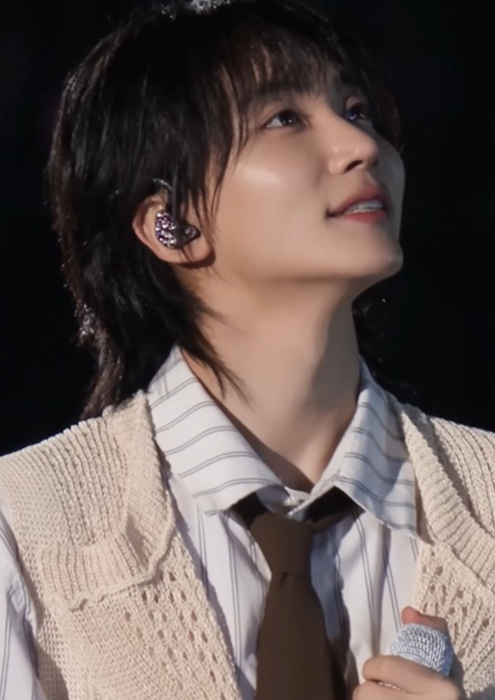 Jeonghan of Seventeen in a cream vest and striped shirt, holding a mic and looking upward during a performance.