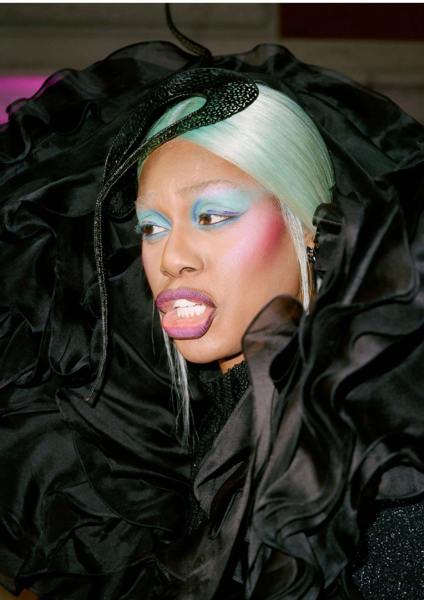 Laverne Cox in a black ruffled outfit with a green headpiece, colorful makeup, and purple lips. (1)