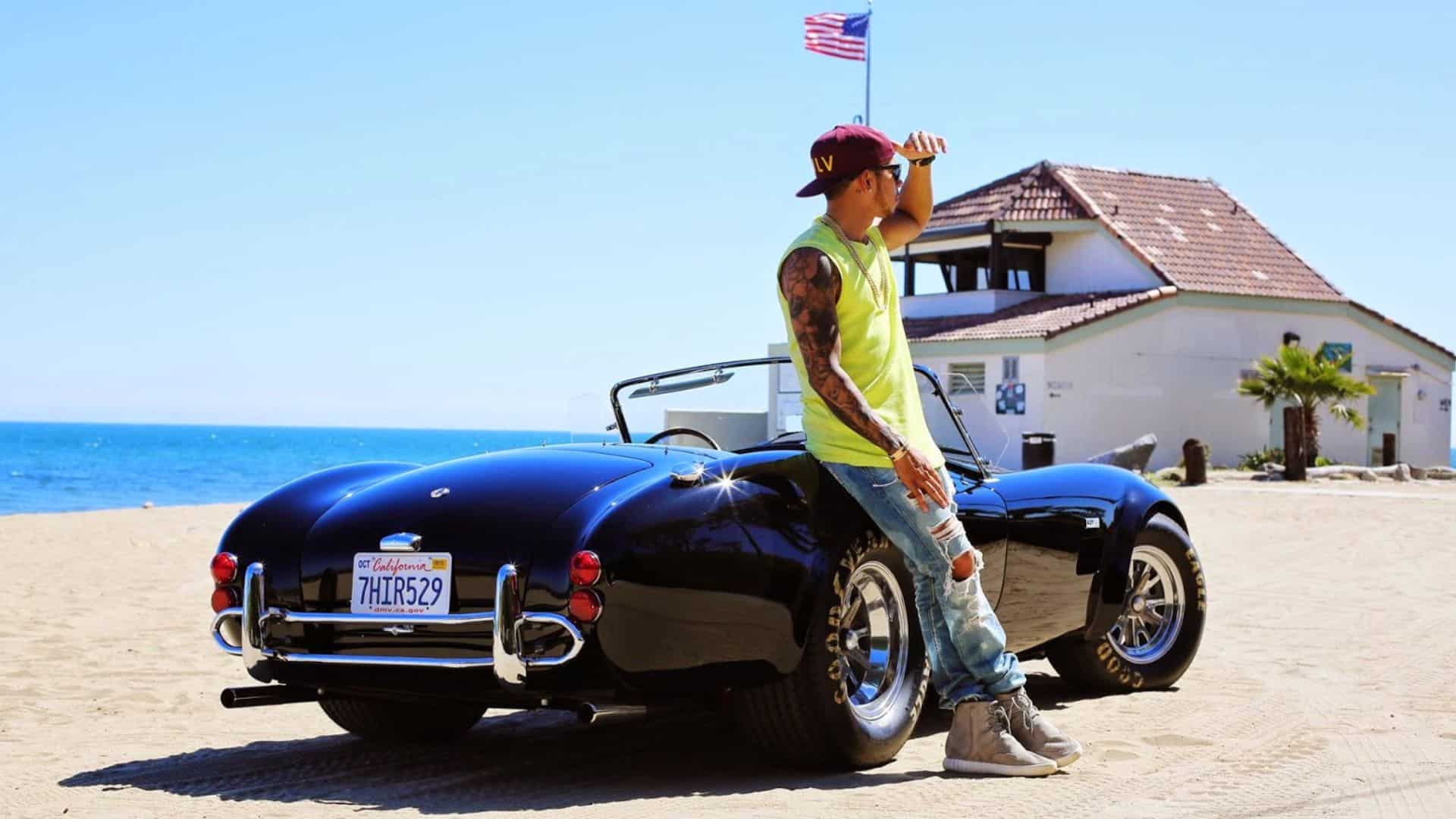 Lewis Hamilton leaning against a classic black convertible sports car near a beachside road on a sunny day.