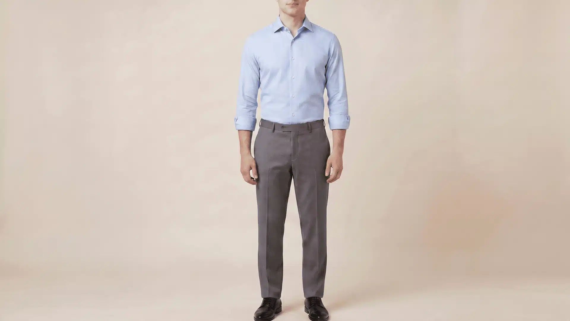 Man wearing a light blue, long-sleeve shirt with charcoal grey pants and black shoes, standing in a neutral background.