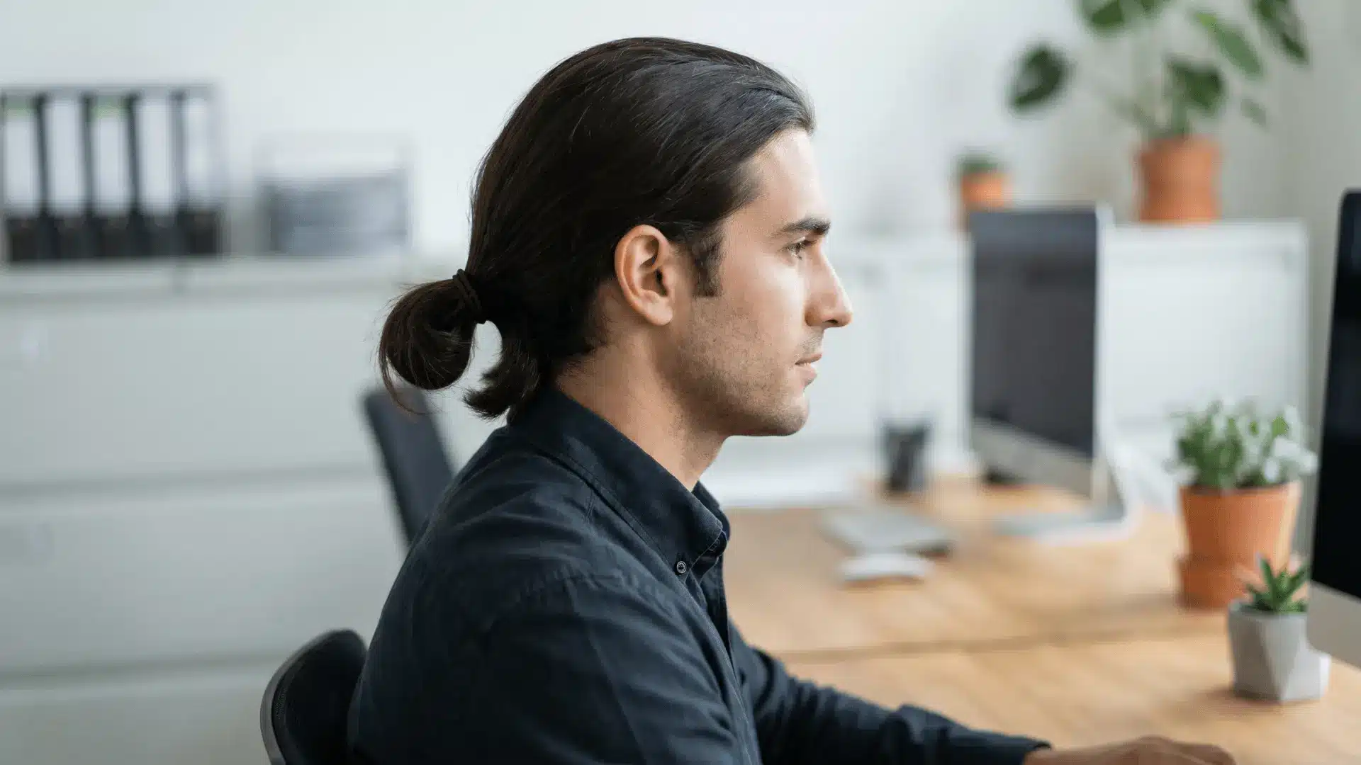 andsome male model in an office with a man bun hairstyle, hair tied into a moderate bun at back.