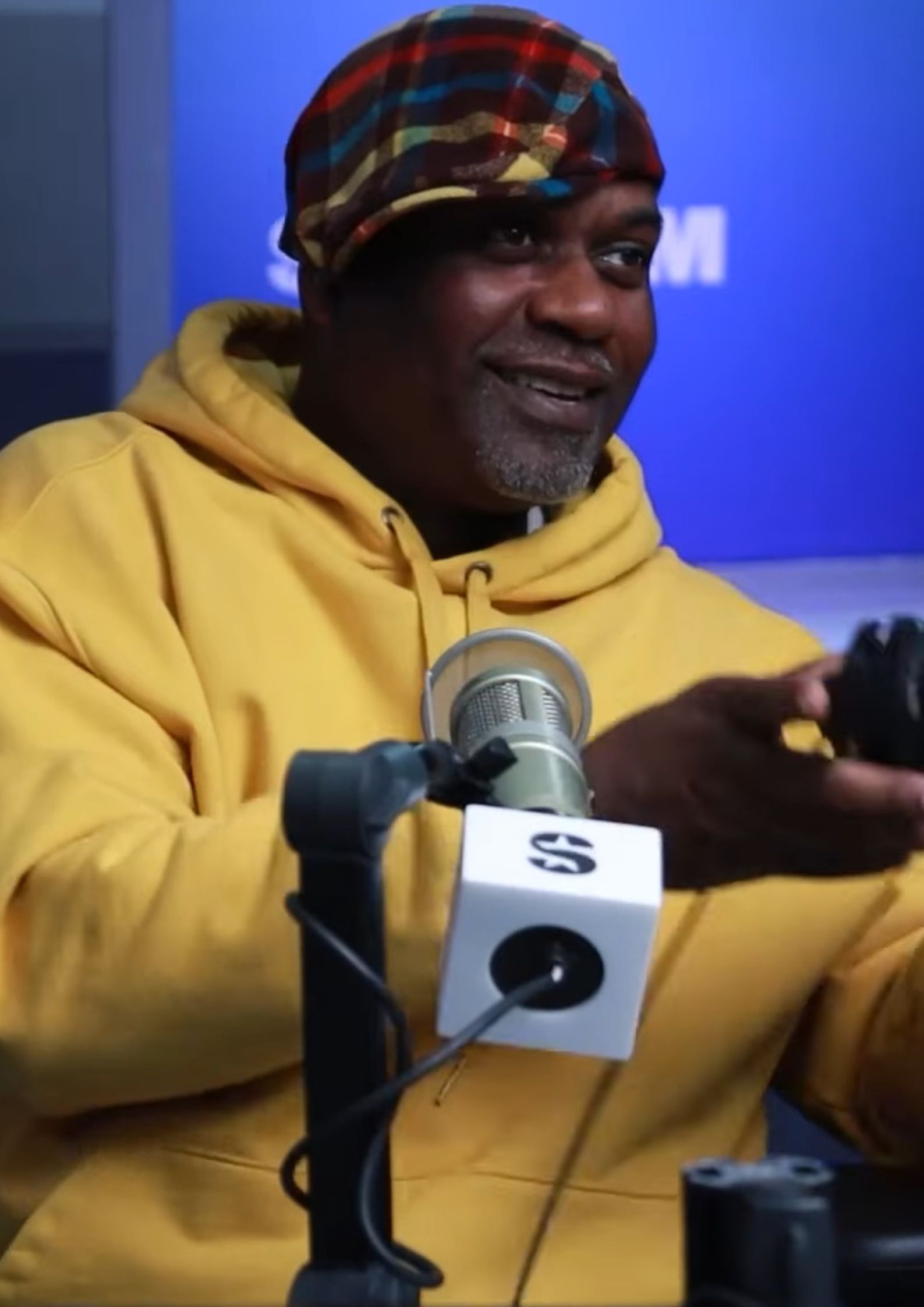 Masta Killa from Wu-Tang Clan wearing a yellow hoodie and multicolored cap, speaking into a microphone during an interview