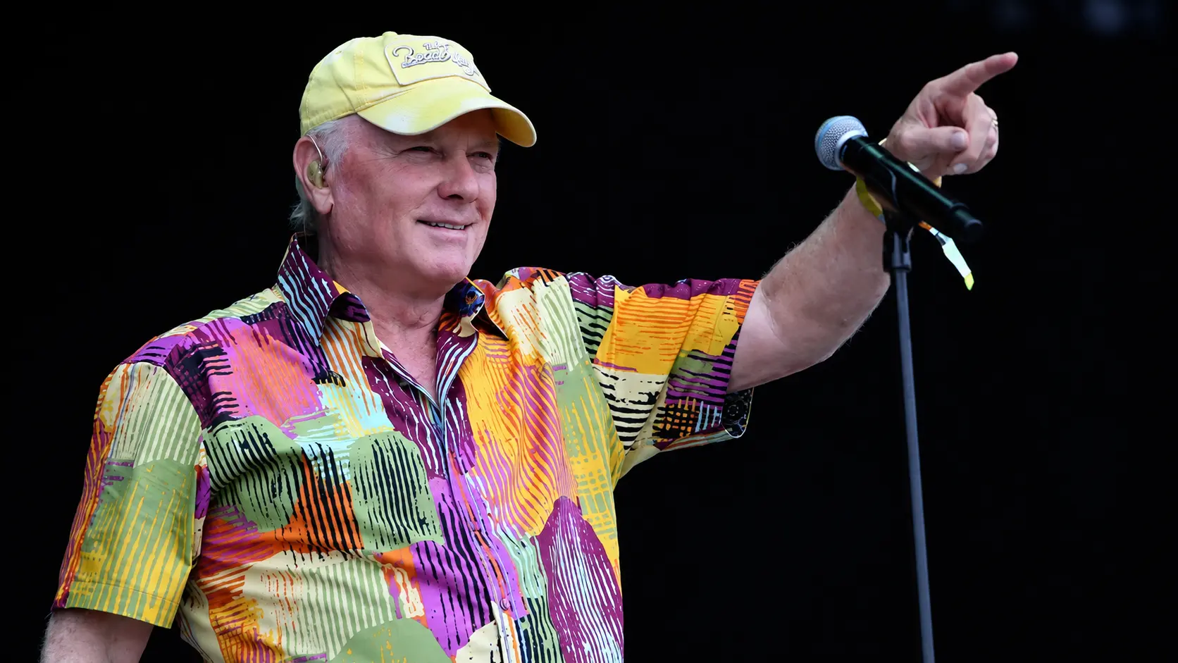 Older man in colorful shirt and yellow cap points while singing on stage with microphone, dark background.