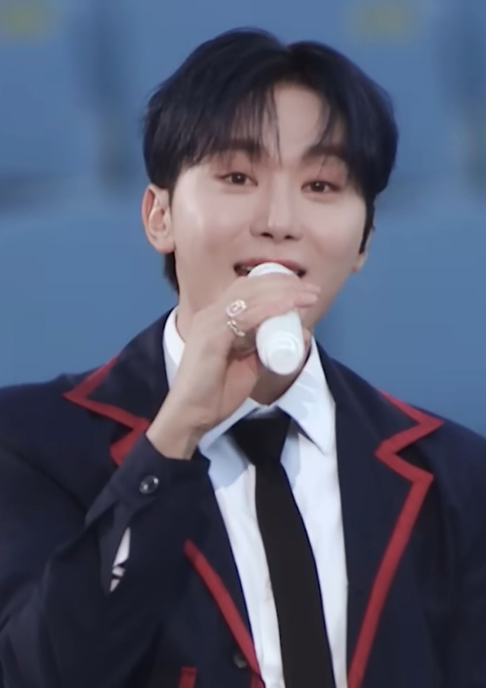 Seventeen member Seungkwan singing on stage, wearing a navy blazer with red trim and a black tie.