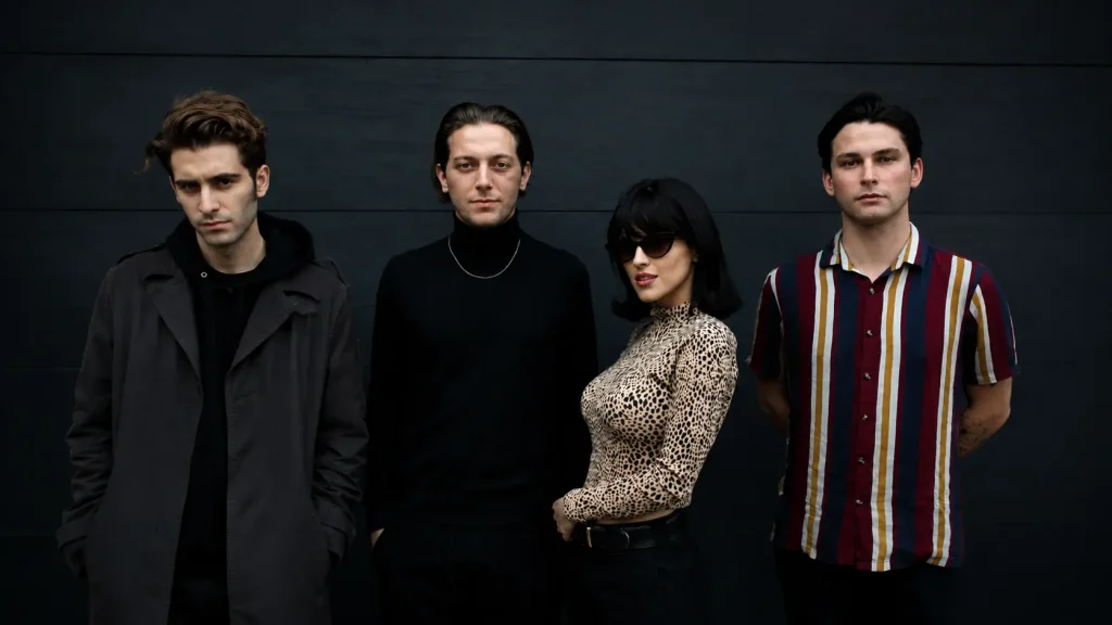 The members of The Marías stand side by side at an event, each showcasing their unique fashion against a dark backdrop
