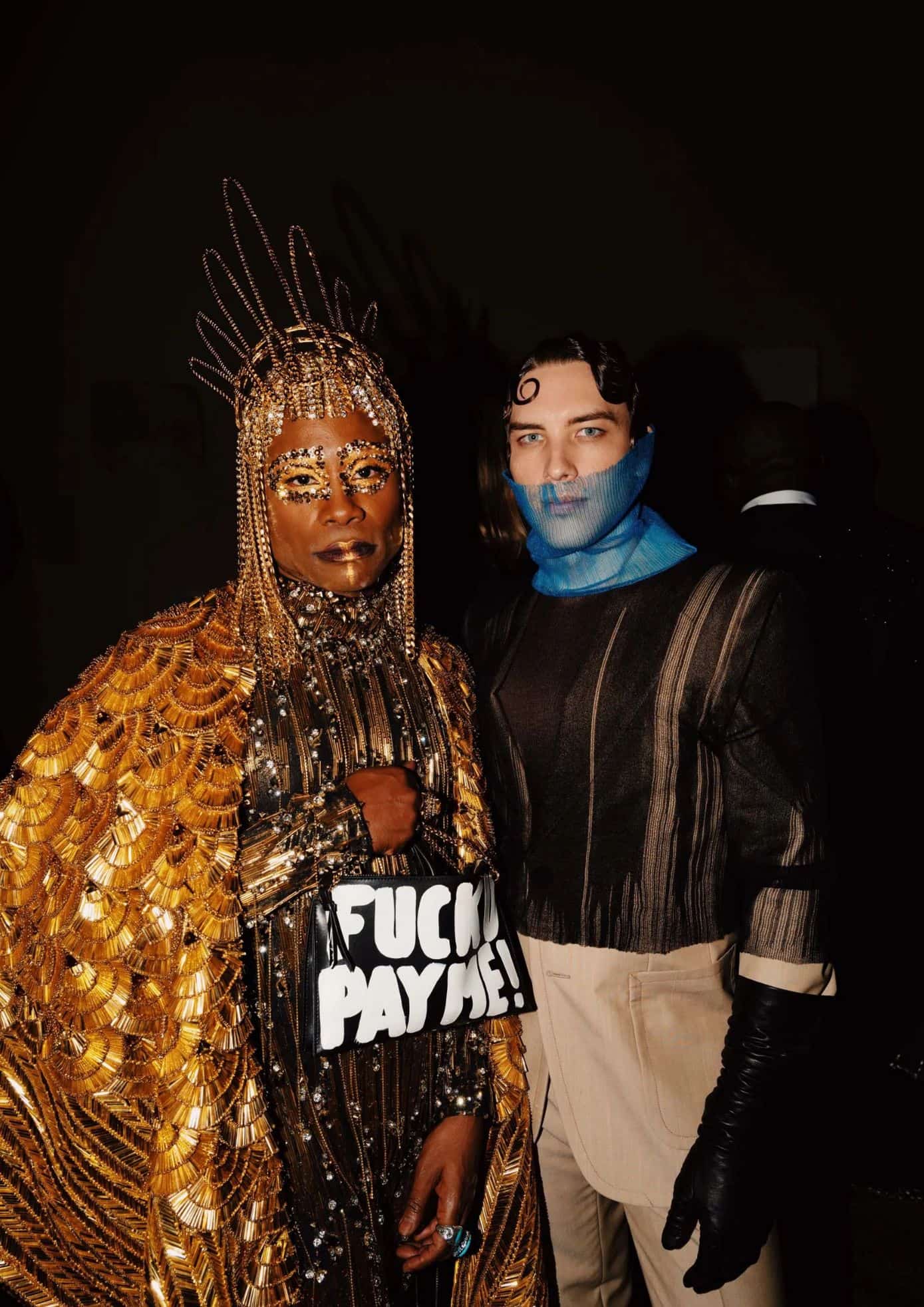 Two guests at the 2019 Met Gala, one in a golden beaded gown with a bag and the other in a sheer outfit with a blue face mask. (1)