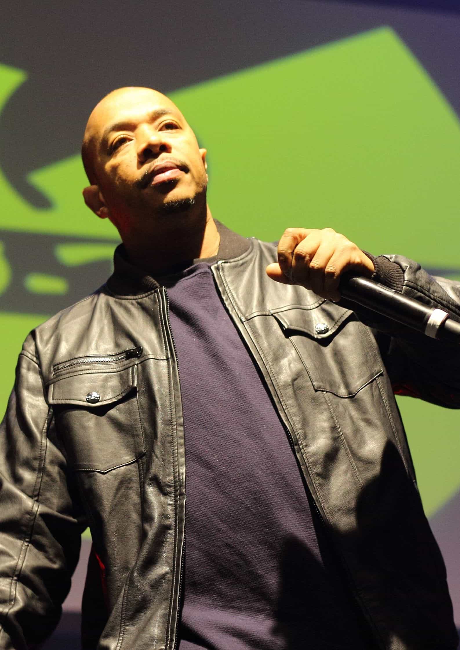 U-God from Wu-Tang Clan wearing a black leather jacket and dark shirt, posing in front of a green background