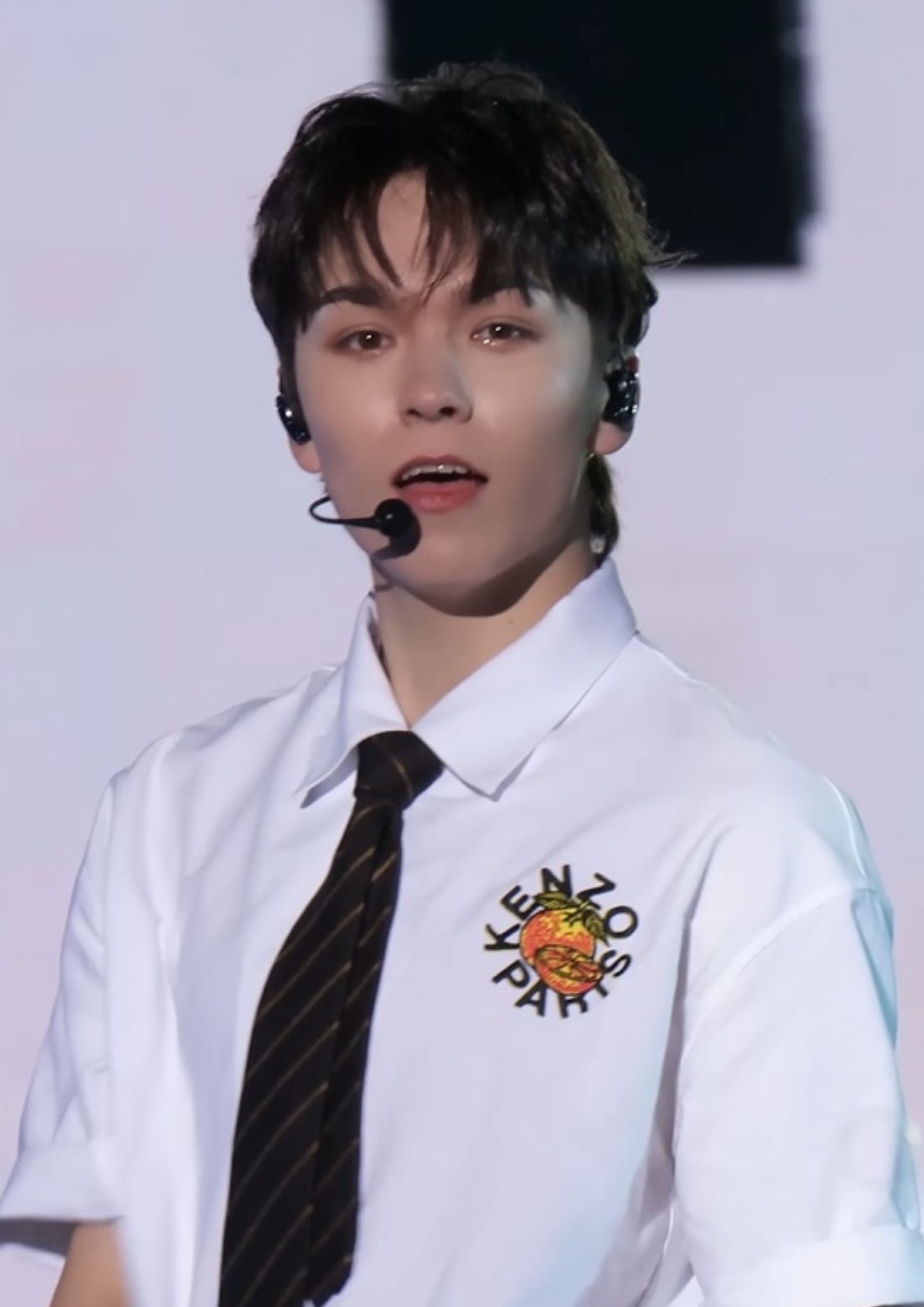 Vernon from Seventeen performing on stage, wearing white shirt, black tie, holding a microphone.